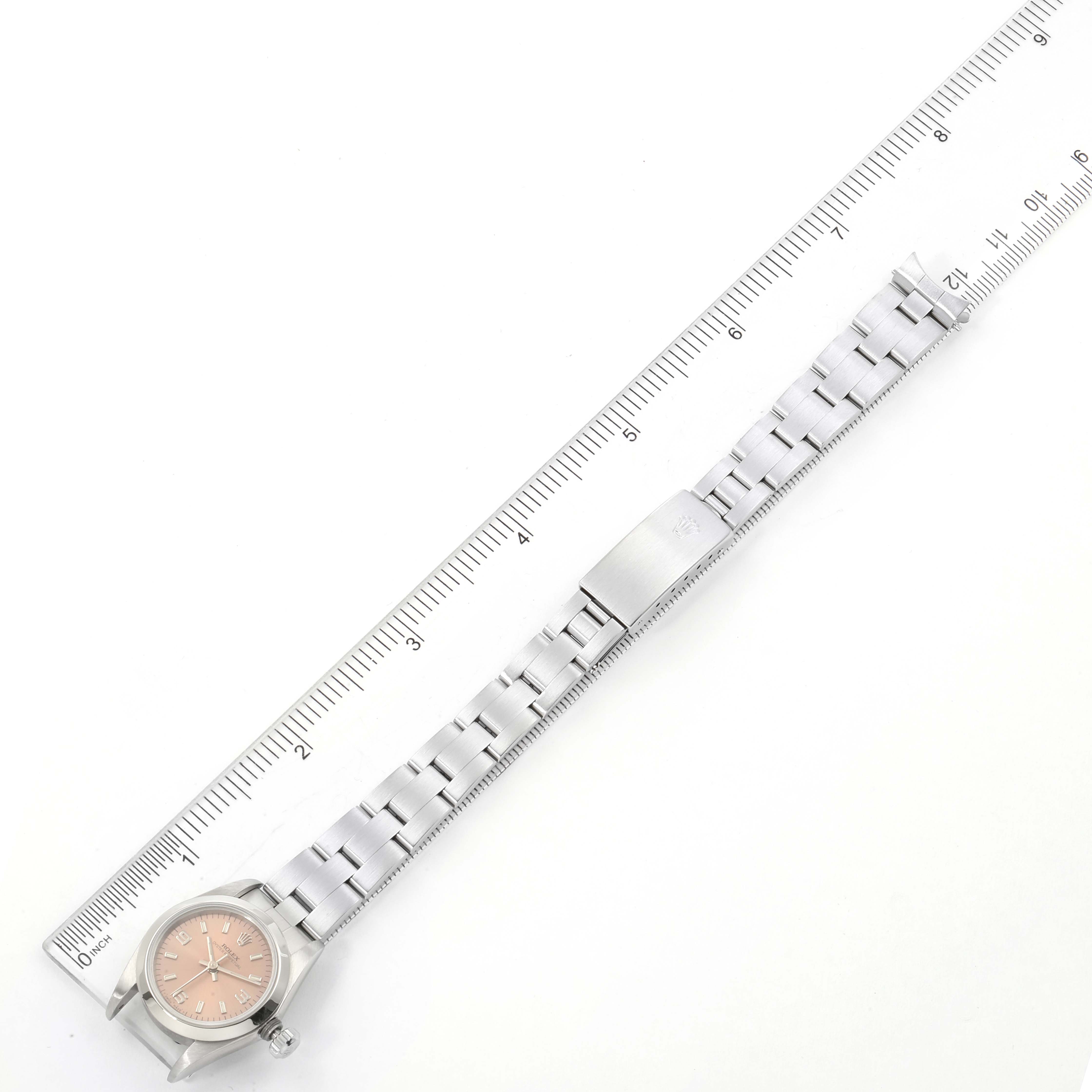 The image shows a top-down view of a Rolex Oyster Perpetual 67180 Women's Stainless Steel Pink Dial 67180 Women's Stainless Steel Pink Dial watch with a full metal bracelet alongside a ruler.