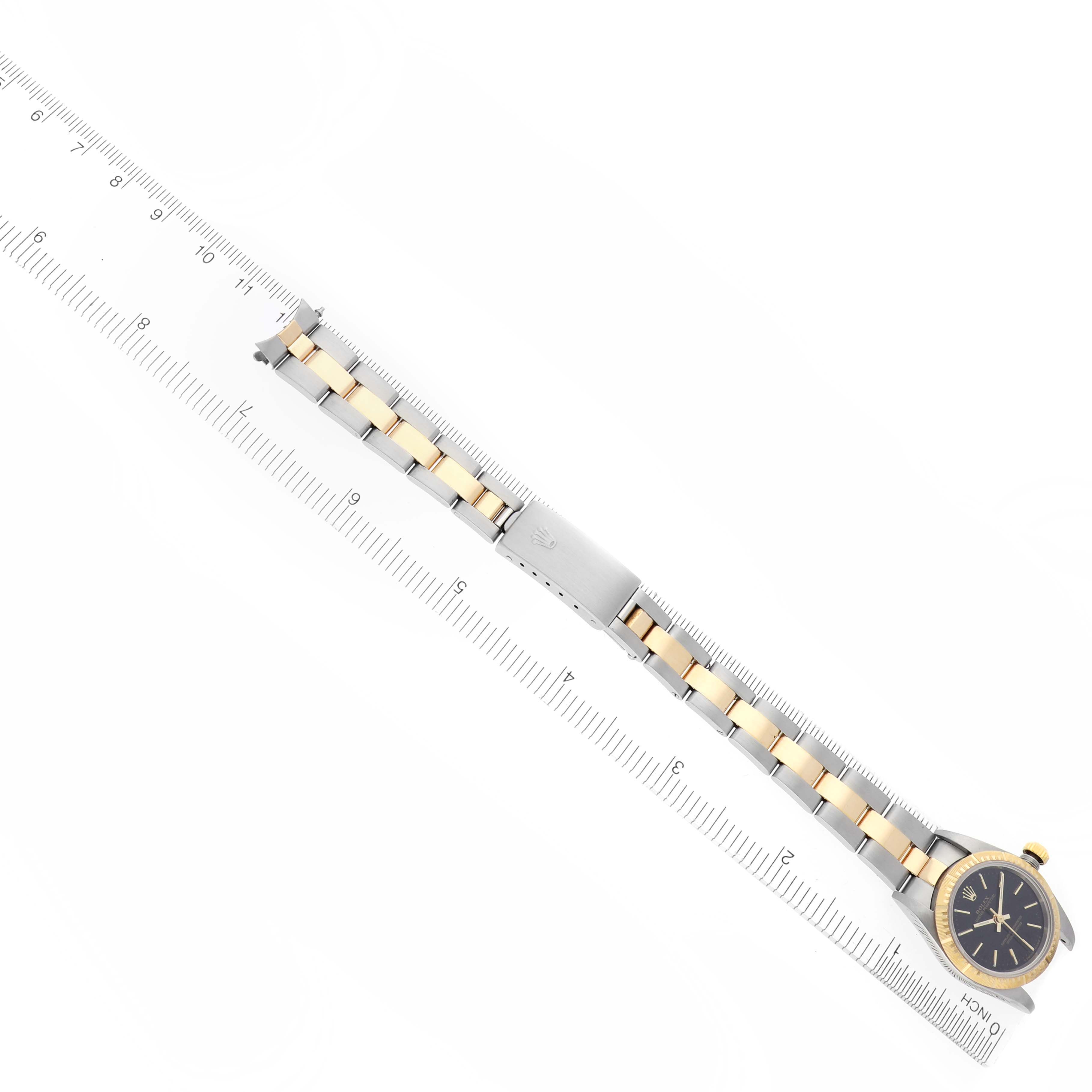 The Rolex Oyster Perpetual 76243 Women's Steel and Gold (two tone) Black Dial 76243 Women's Steel and Gold (two tone) Black Dial watch is shown from an overhead angle, displaying the full bracelet and face alongside a ruler.