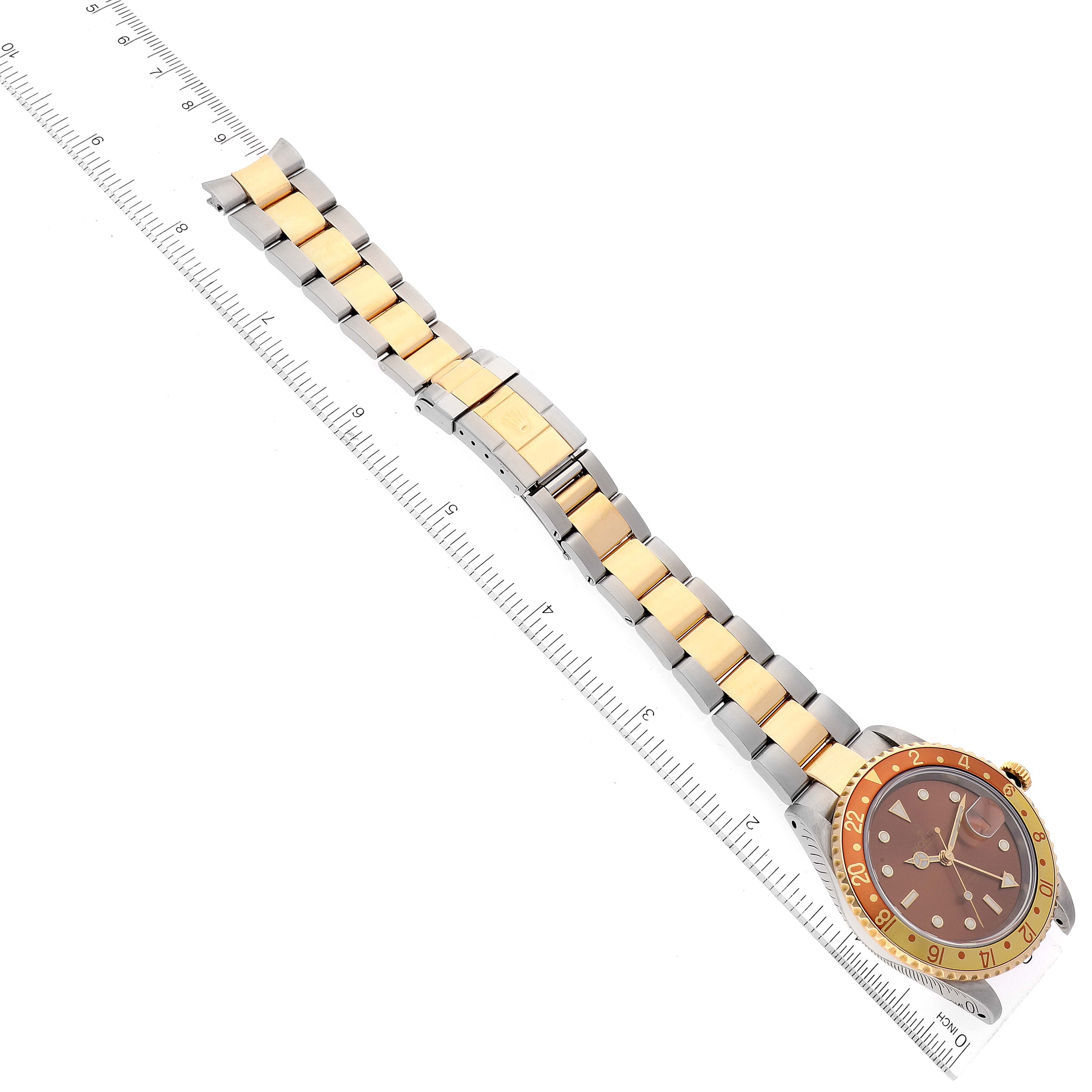 This image shows a Rolex GMT-Master 16713 Men's Steel and Gold (two tone) Brown Dial 16713 Men's Steel and Gold (two tone) Brown Dial watch laid flat beside a ruler, displaying its gold-tone and stainless steel bracelet and dial.