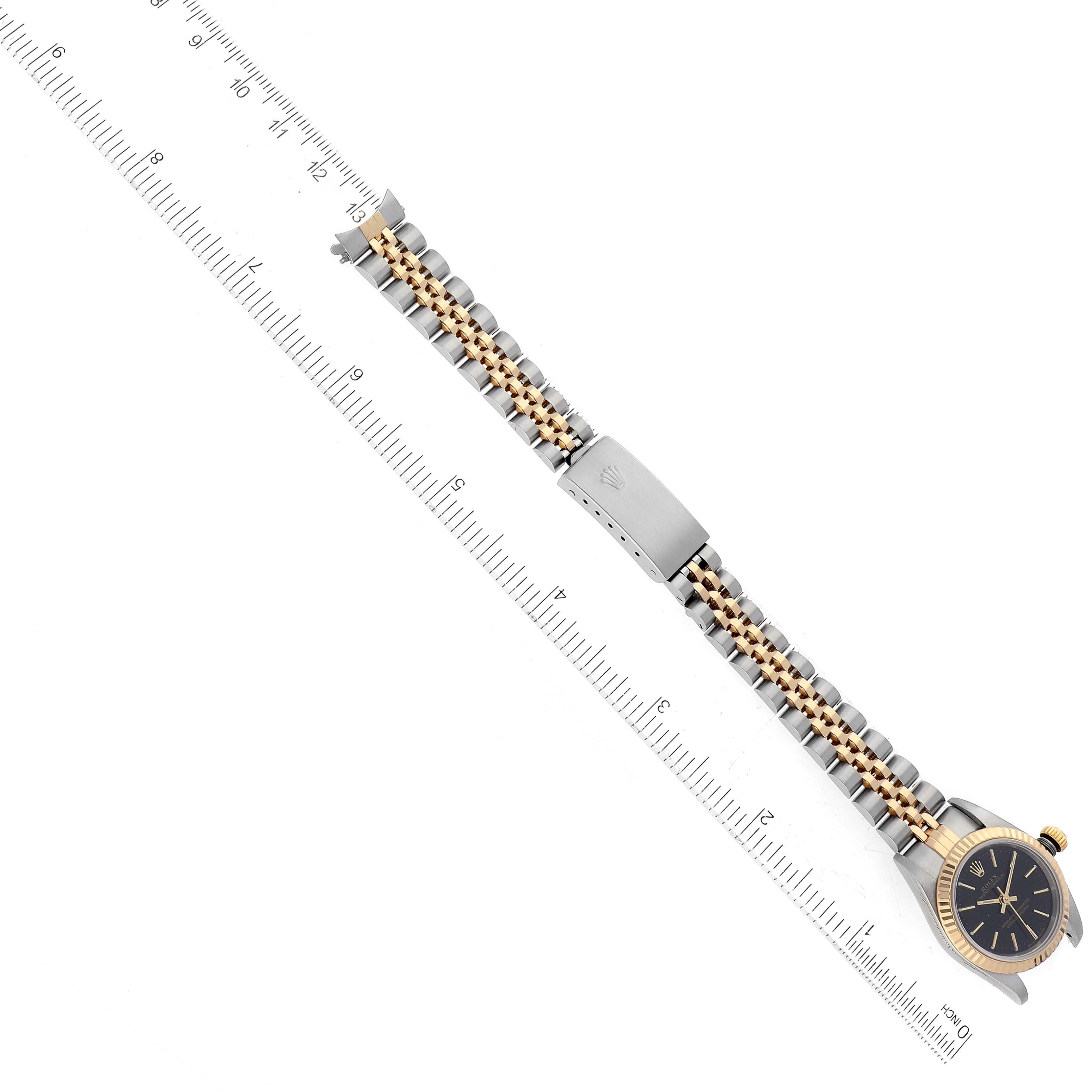 The image shows a top-down view of a Rolex Oyster Perpetual 76193 Women's Steel and Gold (two tone) Black Dial 76193 Women's Steel and Gold (two tone) Black Dial watch and its bracelet alongside a ruler for scale.