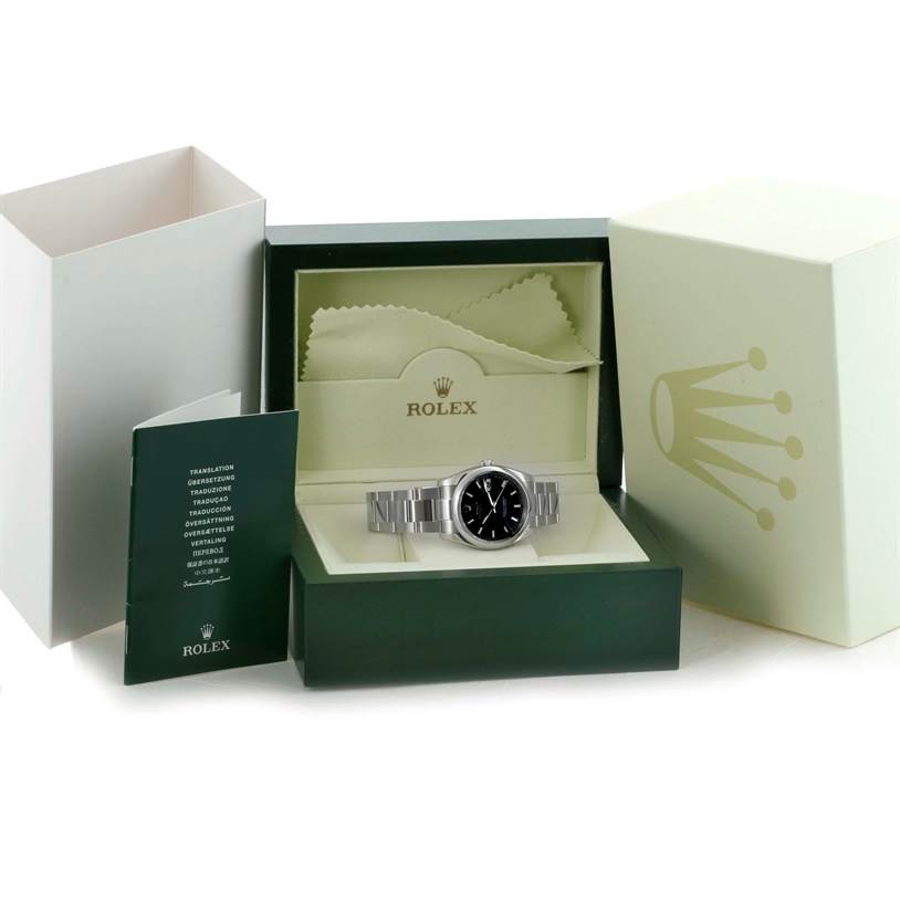 The image shows a Rolex Date 115200 Men's Stainless Steel Black Dial 115200 Men's Stainless Steel Black Dial model watch from the front angle in its box with an accompanying booklet and a cleaning cloth.