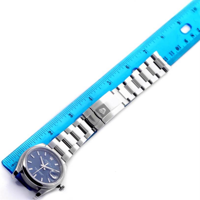 The Rolex Date 115200 Men's Stainless Steel Blue Dial 115200 Men's Stainless Steel Blue Dial model watch is shown from a top-down angle, displaying the face, bracelet, and clasp alongside a ruler.