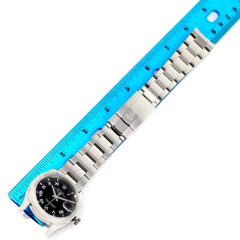 The image shows a top-down view of a Rolex Date 115200 Men's Stainless Steel Black Dial 115200 Men's Stainless Steel Black Dial model watch with a metal bracelet and a blue measuring ruler beside it.