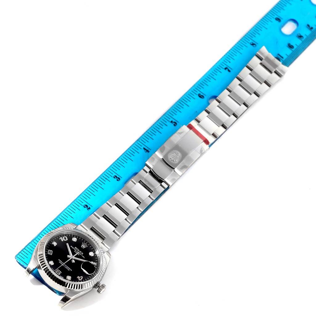 The image shows a top-down angle of a Rolex Date 115234 Men's Stainless Steel Black Dial 115234 Men's Stainless Steel Black Dial watch with its bracelet laid next to a blue ruler.