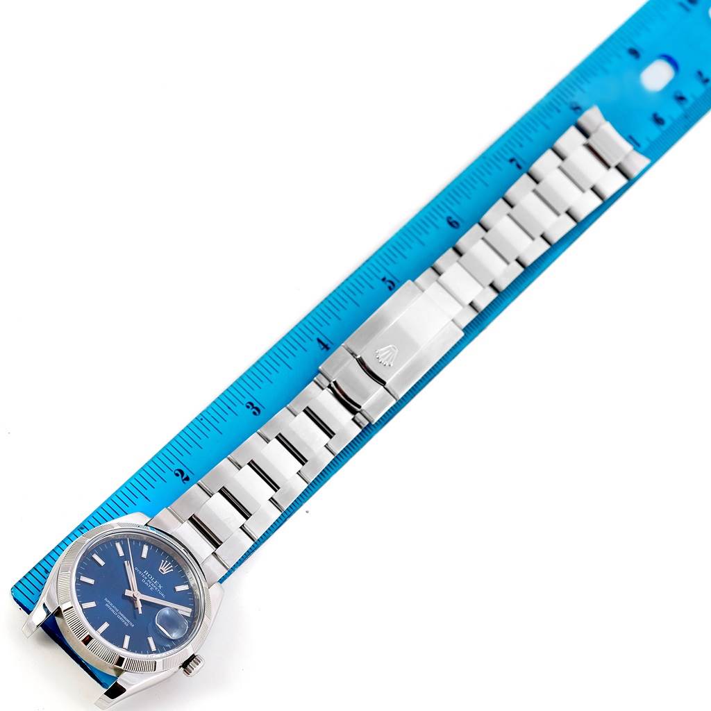 The image shows a top-down view of a Rolex Date 115210 Men's Stainless Steel Blue Dial 115210 Men's Stainless Steel Blue Dial model watch with a blue face and stainless steel bracelet next to a ruler.