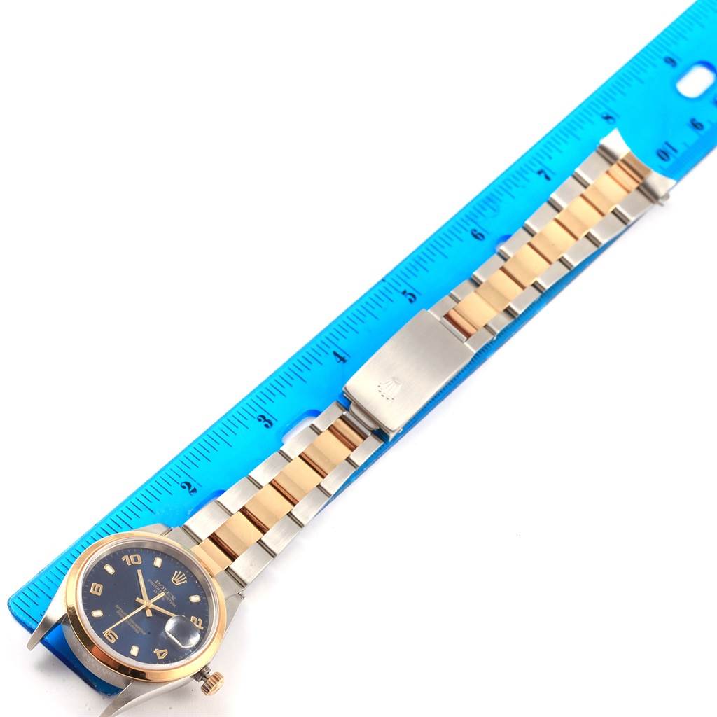 The Rolex Date 15203 Men's Steel and Gold (two tone) Blue Dial 15203 Men's Steel and Gold (two tone) Blue Dial watch is shown from the top view, displaying its face, bezel, bracelet, and a clasp against a blue ruler.