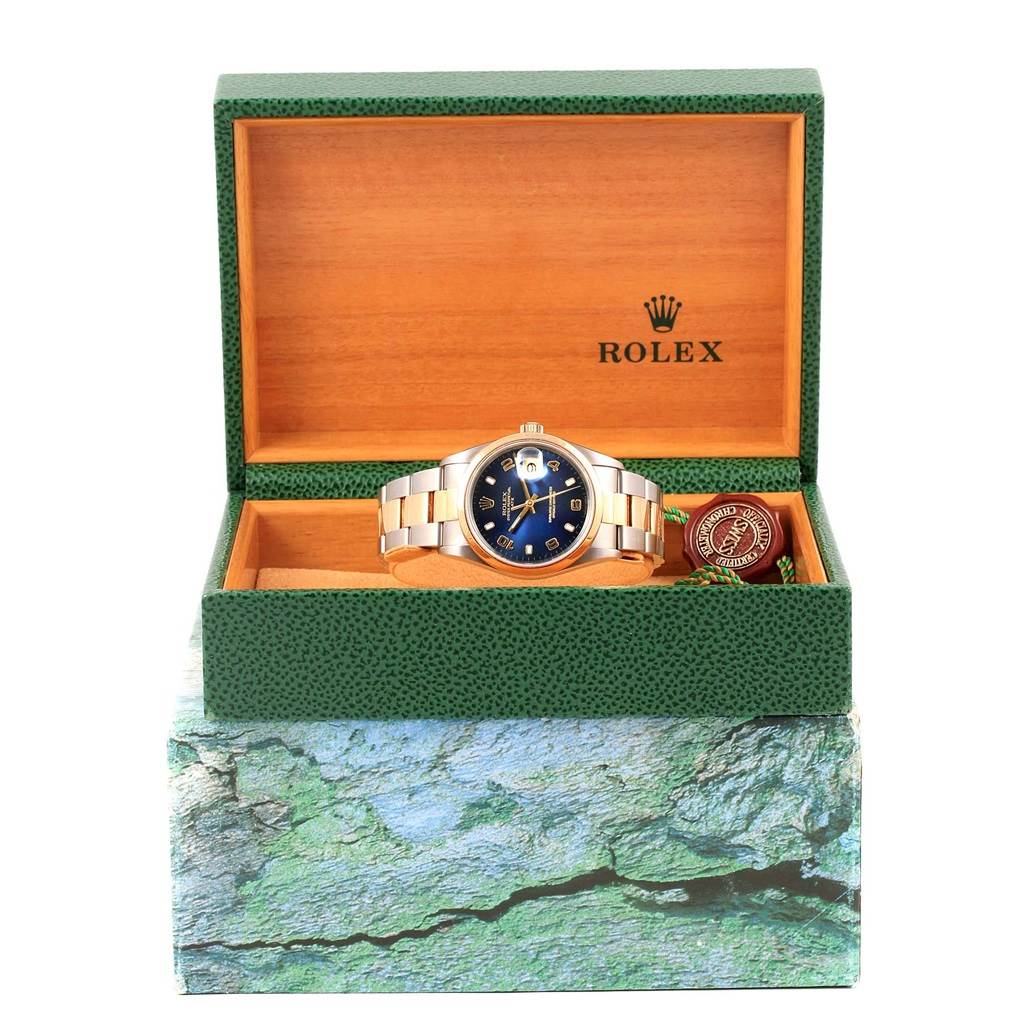 The Rolex Date 15203 Men's Steel and Gold (two tone) Blue Dial 15203 Men's Steel and Gold (two tone) Blue Dial model watch is shown from the front, inside an open green box with additional tags and accessories.