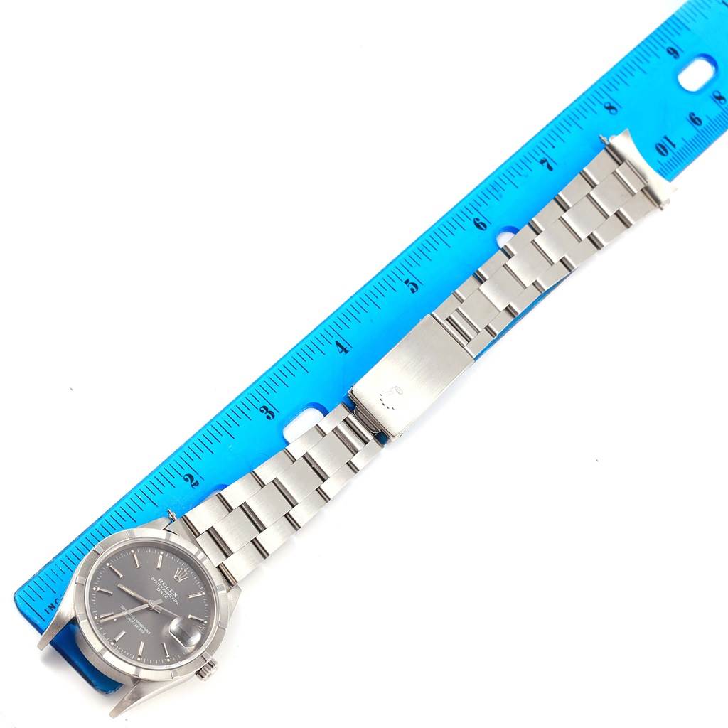 A Rolex Date 15210 Men's Stainless Steel Grey Dial 15210 Men's Stainless Steel Grey Dial model watch is shown laid flat next to a blue ruler, displaying its face, bracelet, and clasp.