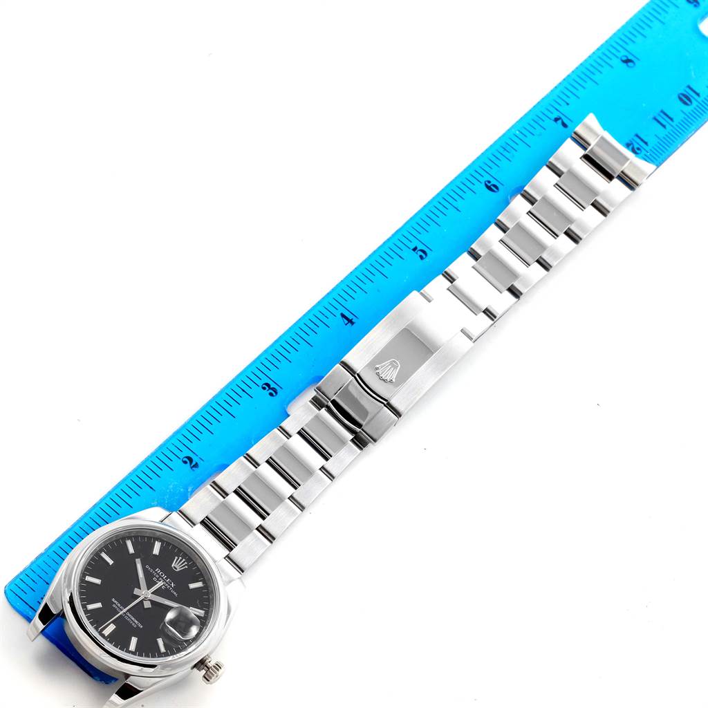 The image shows a Rolex Date 115200 Men's Stainless Steel Black Dial 115200 Men's Stainless Steel Black Dial model watch with a stainless steel band, measuring its length next to a blue ruler.