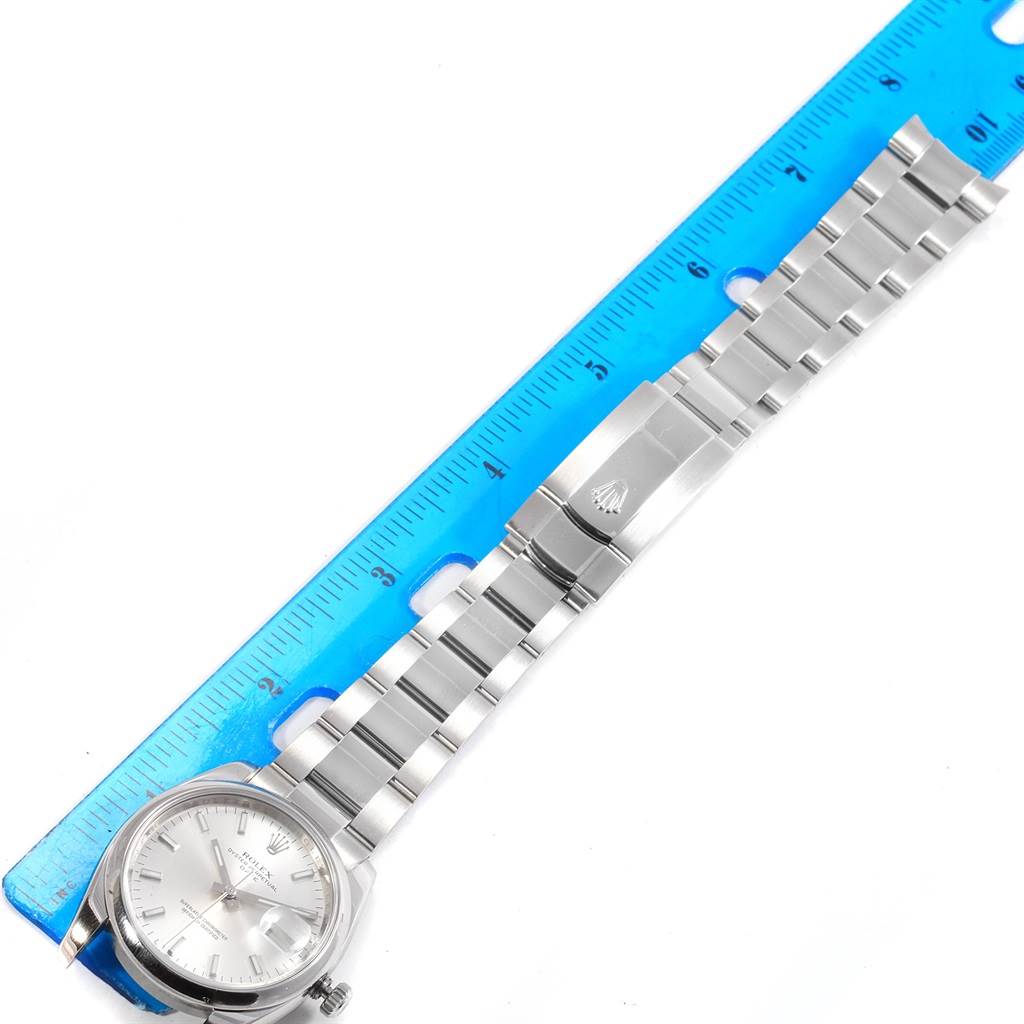 The image shows a side view of a Rolex Date 115200 Men's Stainless Steel Silver Dial 115200 Men's Stainless Steel Silver Dial model watch laying beside a ruler, showcasing the bracelet and clasp.