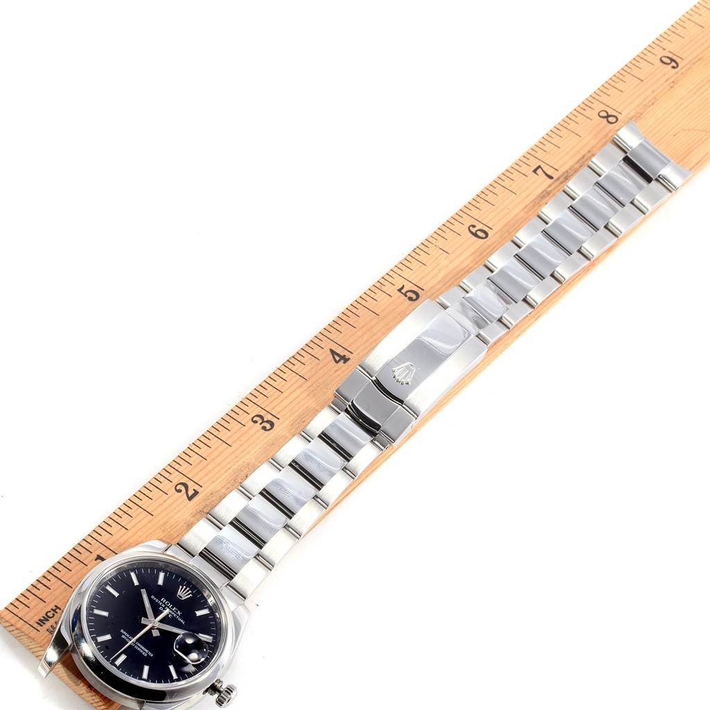 The image shows a top view of a Rolex Date 115200 Men's Stainless Steel Black Dial 115200 Men's Stainless Steel Black Dial model watch, including its face, bracelet, and clasp, with a ruler for size reference.