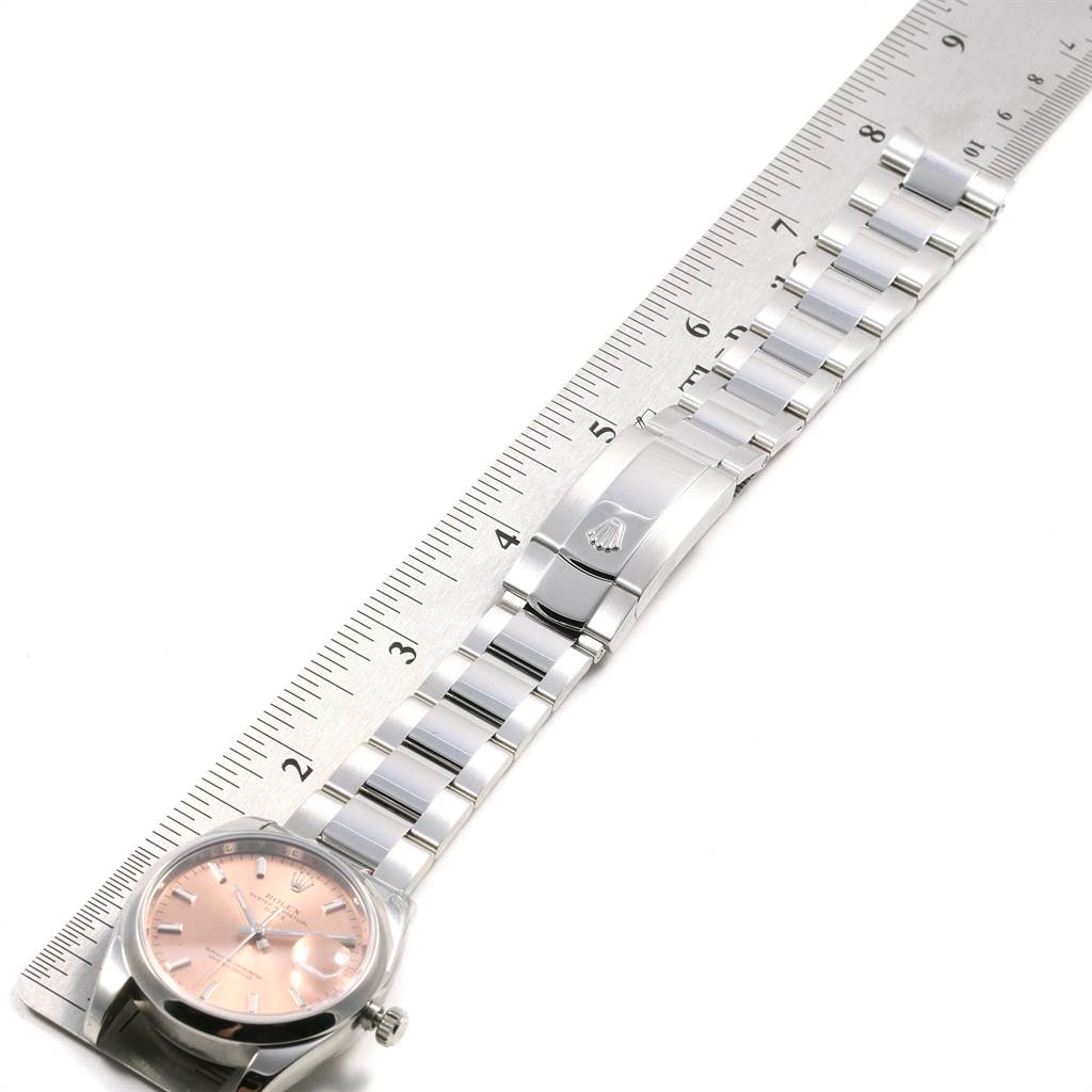 The image shows a Rolex Date 115200 Men's Stainless Steel Pink Dial 115200 Men's Stainless Steel Pink Dial model watch laid flat alongside a ruler, highlighting the dial and bracelet.