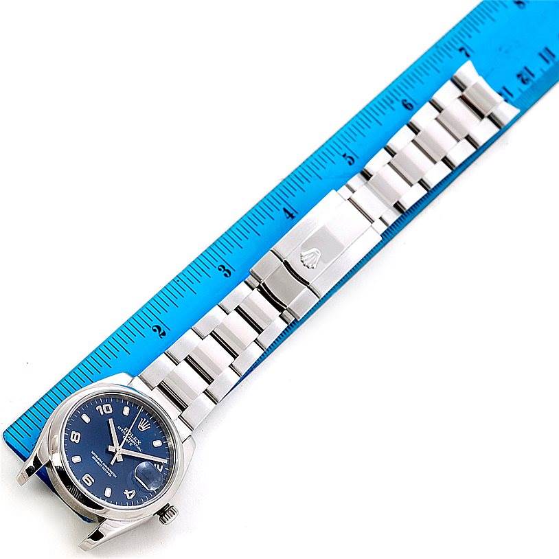 The image shows a Rolex Date 115200 Men's Stainless Steel Blue Dial 115200 Men's Stainless Steel Blue Dial model watch laid flat next to a ruler, highlighting the watch face and full bracelet.