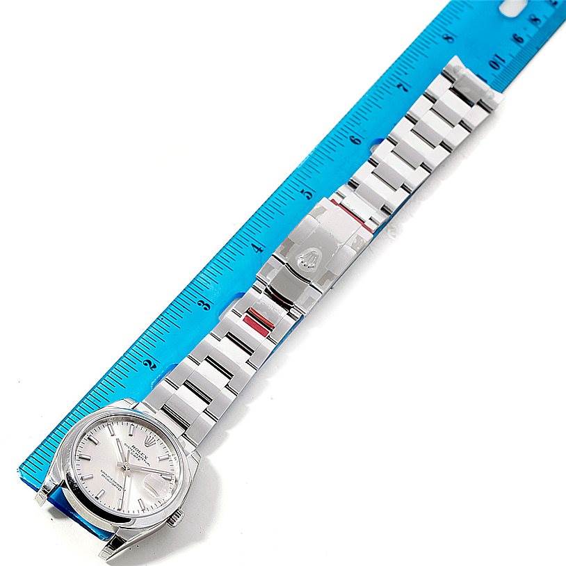 The Rolex Date 115200 Men's Stainless Steel Silver Dial 115200 Men's Stainless Steel Silver Dial model watch is shown flat with the face-up and the metal bracelet extended beside a blue ruler.