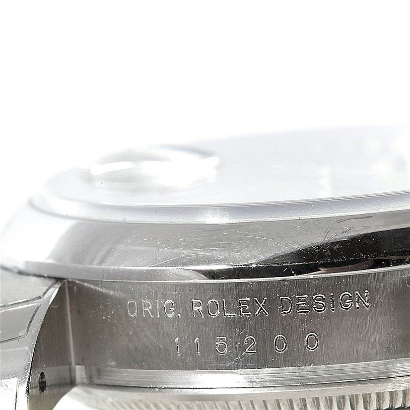 The image shows a close-up of the side of a Rolex Date 115200 Men's Stainless Steel Silver Dial 115200 Men's Stainless Steel Silver Dial model watch, featuring the "Orig. Rolex Design" inscription and model number 115200.
