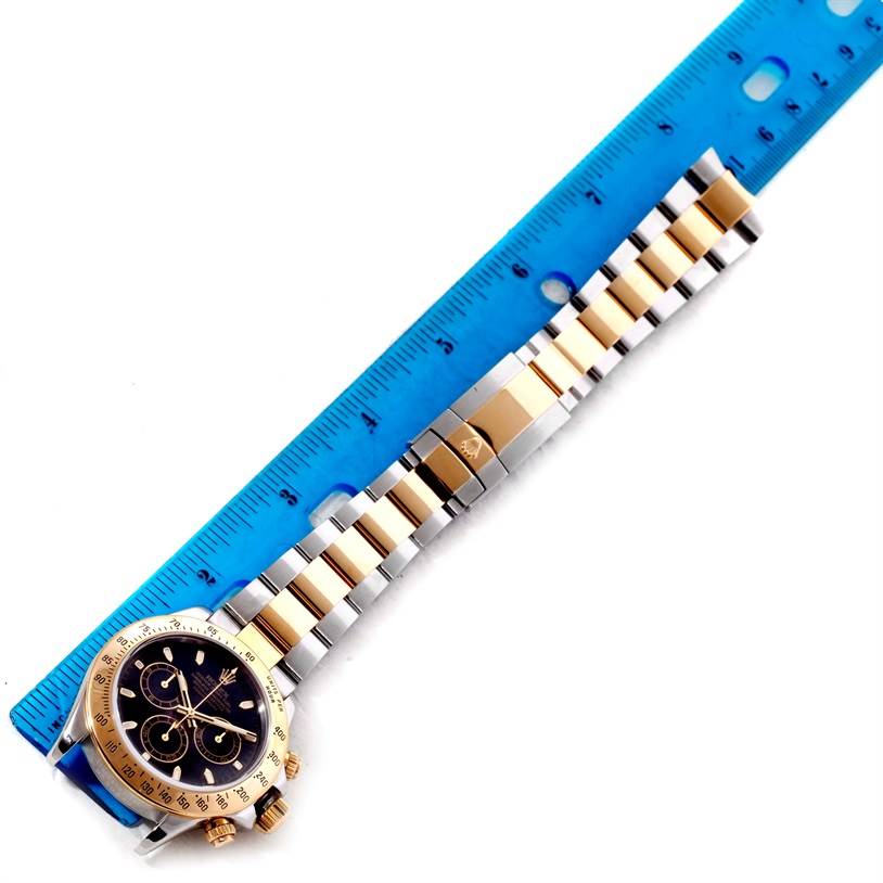 The Rolex Daytona 116523 Men's Steel and Gold (two tone) Black Dial 116523 Men's Steel and Gold (two tone) Black Dial watch is shown flat, with a stainless steel and gold bracelet alongside a blue ruler for scale.