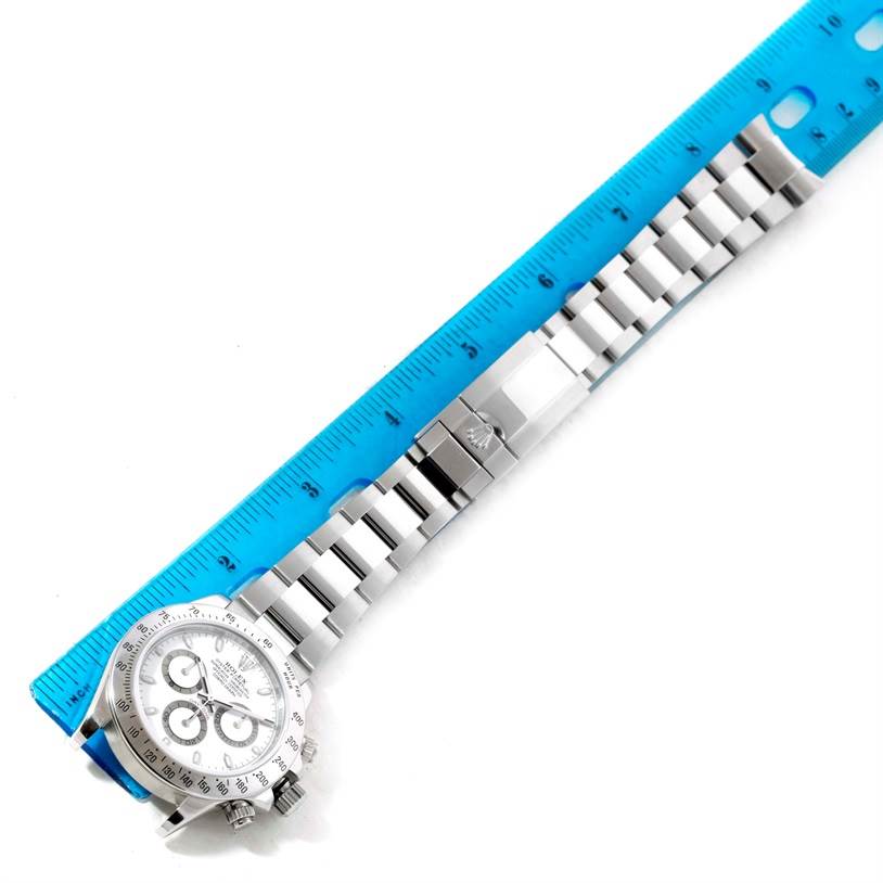 The Rolex Daytona 116520 Men's Stainless Steel White Dial 116520 Men's Stainless Steel White Dial watch is shown from an overhead angle alongside a blue measuring ruler, highlighting its bracelet and clasp.