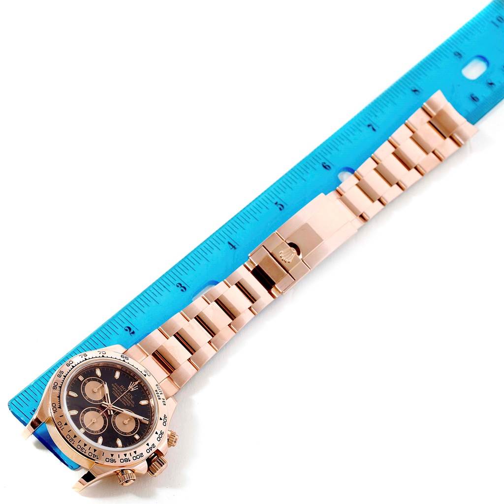 The image shows an overhead view of a Rolex Daytona 116505 Men's Rose Gold Black Dial 116505 Men's Rose Gold Black Dial watch next to a blue ruler, highlighting its length and bracelet.