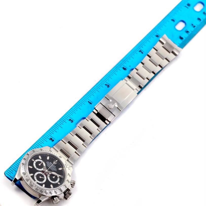 The image shows a top view of a Rolex Daytona 116520 Men
s Stainless Steel Black Dial 116520 Men
s Stainless Steel Black Dial watch, including the face, bezel, and metal band alongside a blue ruler.