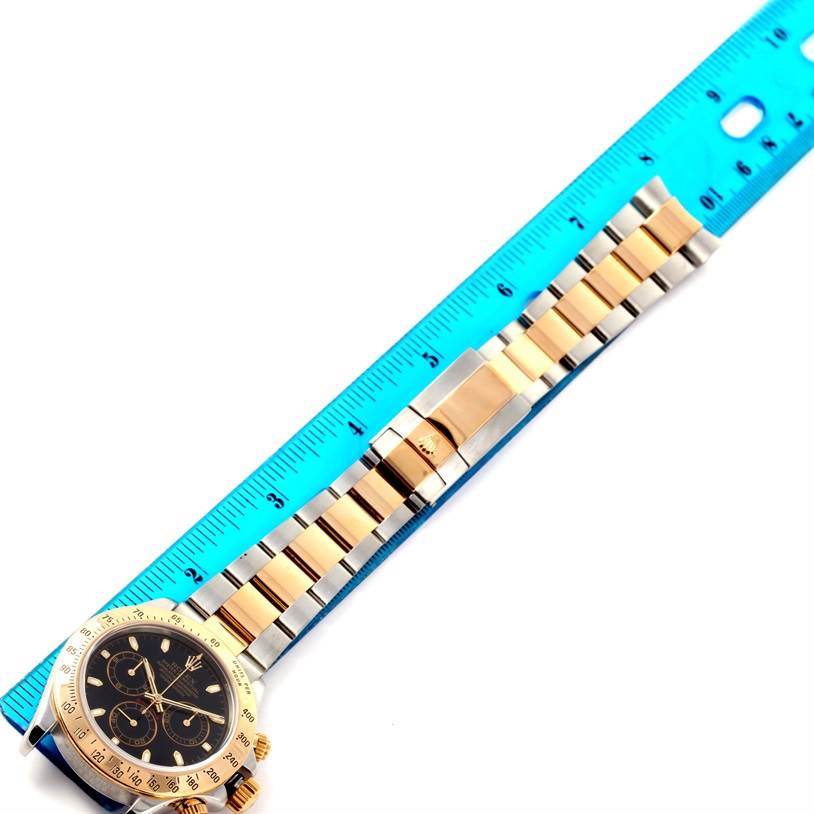 The image shows a Rolex Daytona 116523 Men's Steel and Gold (two tone) Black Dial 116523 Men's Steel and Gold (two tone) Black Dial watch placed flat against a ruler, highlighting the bezel, face, and bracelet in gold and stainless steel.