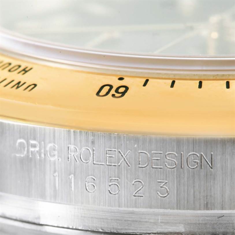 The image shows a close-up of the bezel and case side of a Rolex Daytona 116523 Men's Steel and Gold (two tone) Black Dial 116523 Men's Steel and Gold (two tone) Black Dial watch, featuring engraved text and part of the tachymeter scale.