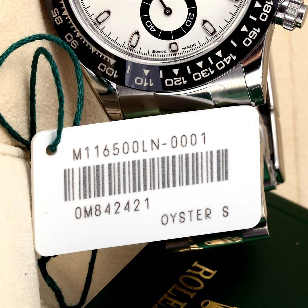 The image shows an angled close-up of the Rolex Daytona 116500 Men's Stainless Steel White Dial 116500 Men's Stainless Steel White Dial watch face, bezel, and the attached tag with model and serial numbers.