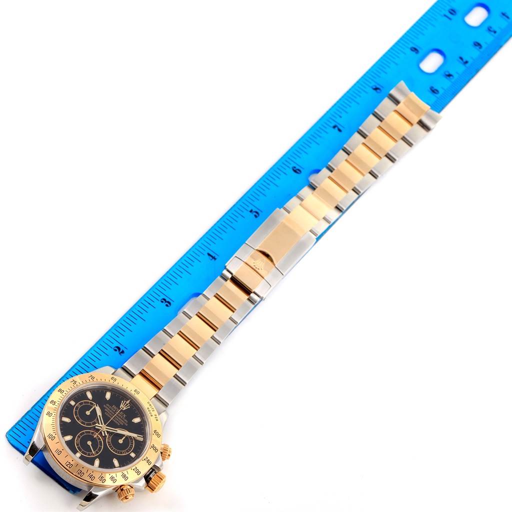 The image shows a Rolex Daytona 116523 Men's Steel and Gold (two tone) Black Dial 116523 Men's Steel and Gold (two tone) Black Dial watch with a gold and silver bracelet next to a blue ruler, taken from an overhead angle.