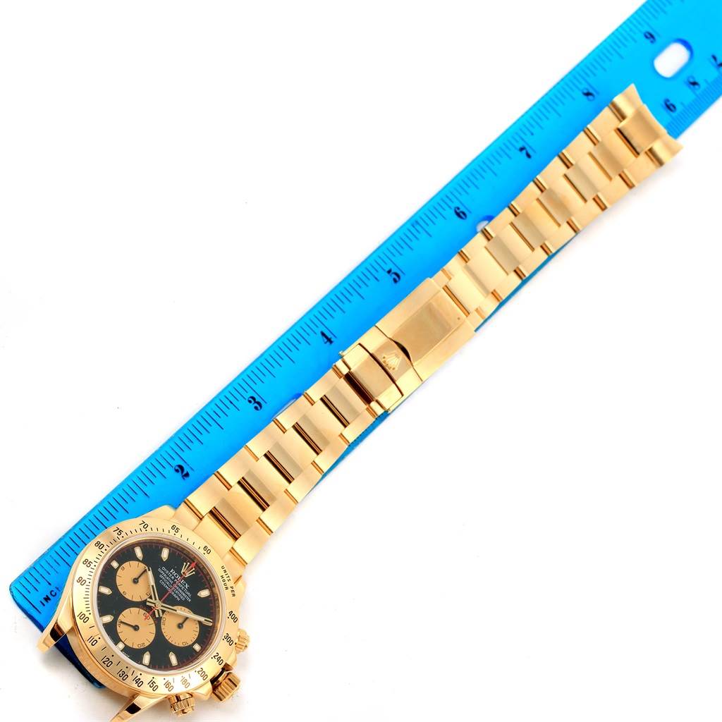 The image shows a gold Rolex Daytona 116528 Men's Yellow Gold Black Dial 116528 Men's Yellow Gold Black Dial watch laid flat next to a blue ruler, displaying the watch's face and bracelet.