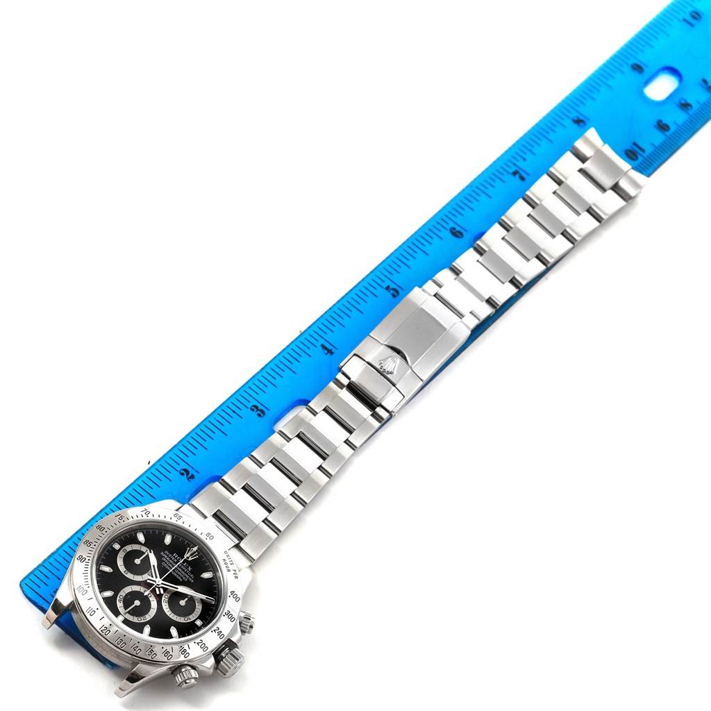 The image shows a Rolex Daytona 116520 Men's Stainless Steel Black Dial 116520 Men's Stainless Steel Black Dial watch laid flat next to a blue ruler, displaying the dial and silver bracelet.