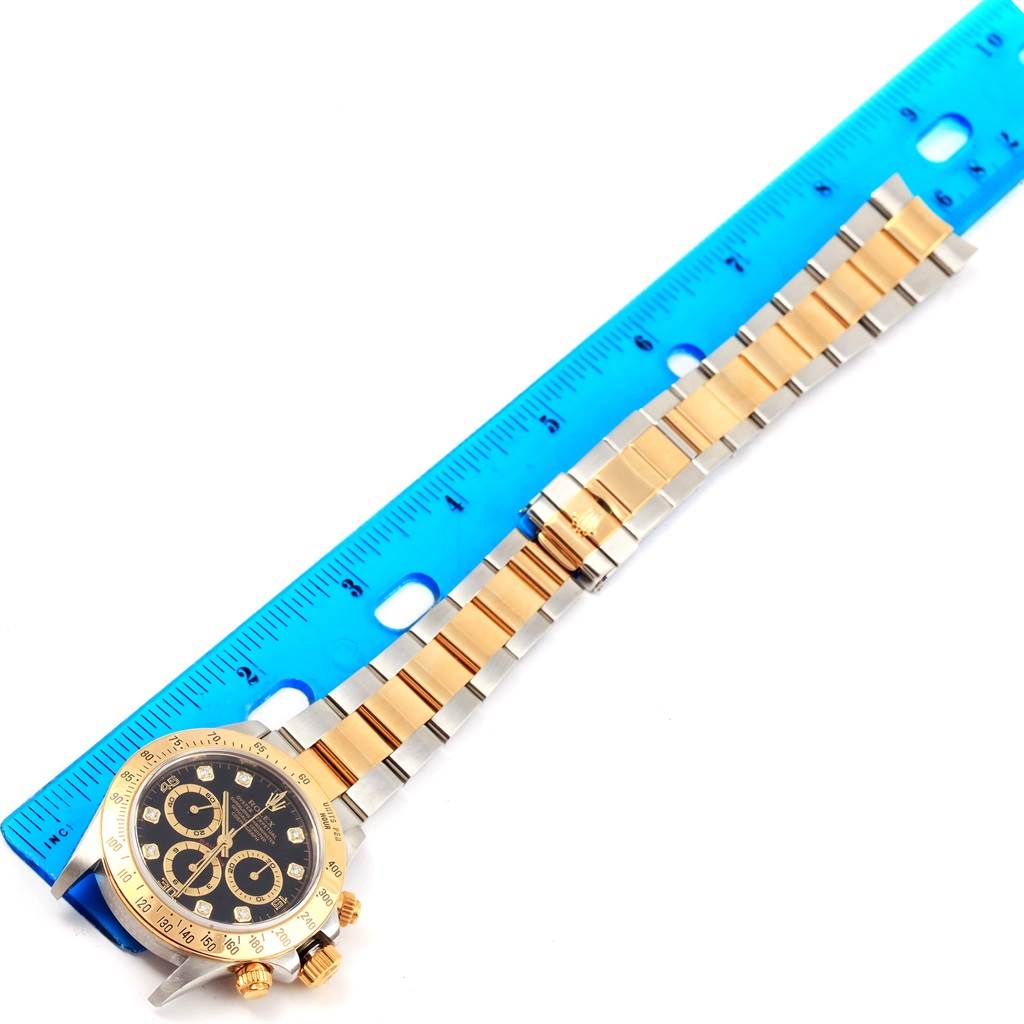 The image shows a Rolex Daytona 16523 Men's Steel and Gold (two tone) Black Dial 16523 Men's Steel and Gold (two tone) Black Dial watch laid flat beside a blue ruler, highlighting its bracelet, case, and dial.