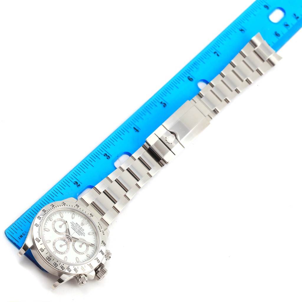 The image shows a Rolex Daytona 116520 Men
s Stainless Steel White Dial 116520 Men
s Stainless Steel White Dial watch with a full view of the face, crown, and metal bracelet, placed next to a ruler.