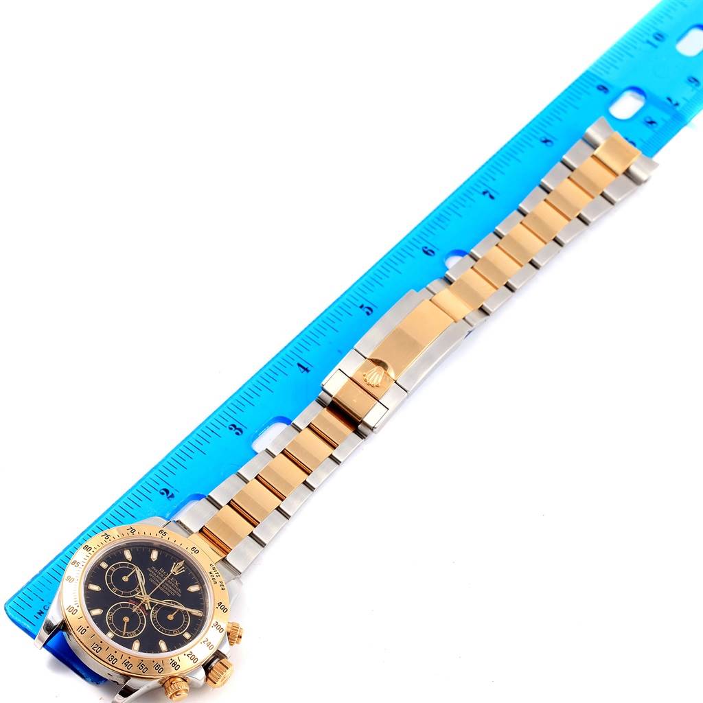 This image shows a top-down view of a Rolex Daytona 116523 Men's Steel and Gold (two tone) Black Dial 116523 Men's Steel and Gold (two tone) Black Dial watch, including the face, bezel, and bracelet positioned next to a blue ruler.