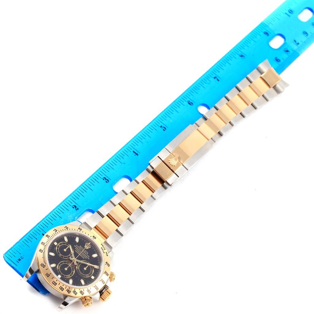 This image shows a top view of a Rolex Daytona 116523 Men's Steel and Gold (two tone) Black Dial 116523 Men's Steel and Gold (two tone) Black Dial watch with its full bracelet laid flat beside a blue ruler for size comparison.