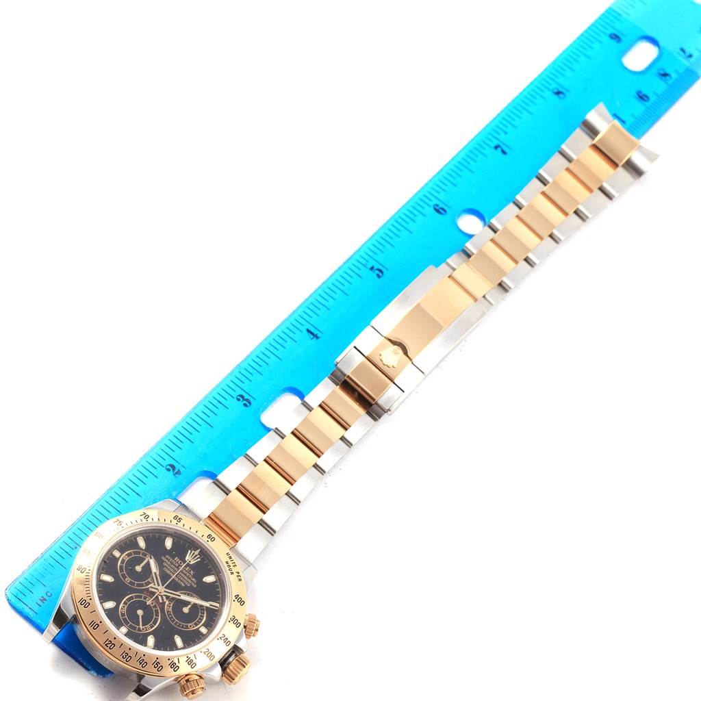 This Rolex Daytona 116523 Men's Steel and Gold (two tone) Black Dial 116523 Men's Steel and Gold (two tone) Black Dial watch is photographed flat alongside a ruler, showing the dial, bezel, and bracelet.