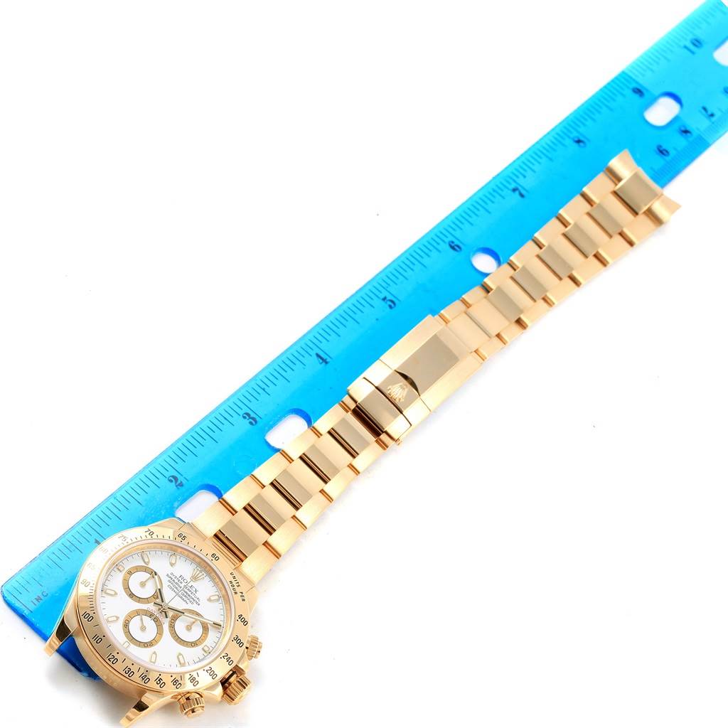 This image shows a Rolex Daytona 116528 Men's Yellow Gold White Dial 116528 Men's Yellow Gold White Dial watch laid flat beside a blue ruler, displaying its face, band, and clasp.
