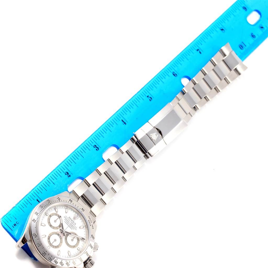 The image shows a Rolex Daytona 116520 Men
s Stainless Steel White Dial 116520 Men
s Stainless Steel White Dial watch laid flat with its bracelet next to a blue ruler for scale.