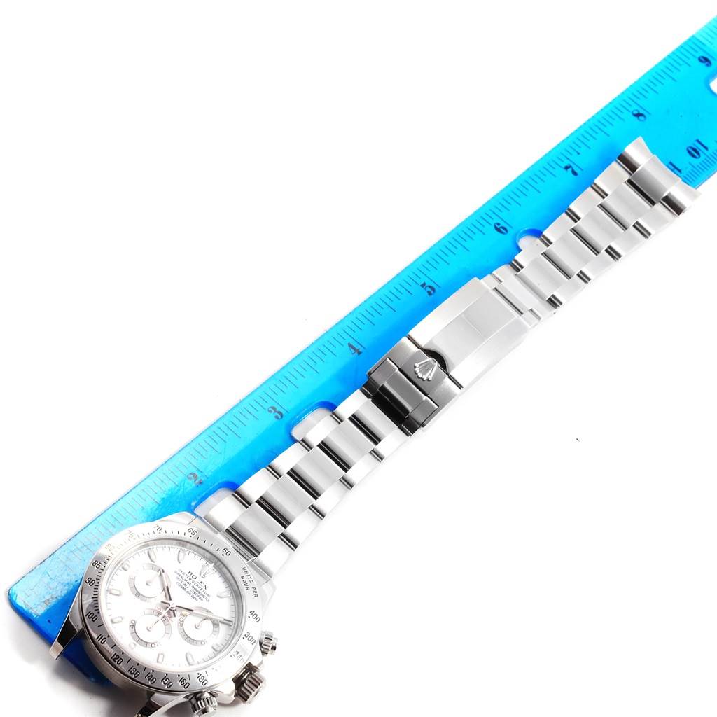 The image shows an overhead view of a Rolex Daytona 116520 Men's Stainless Steel White Dial 116520 Men's Stainless Steel White Dial watch lying next to a blue ruler, displaying the face and bracelet.