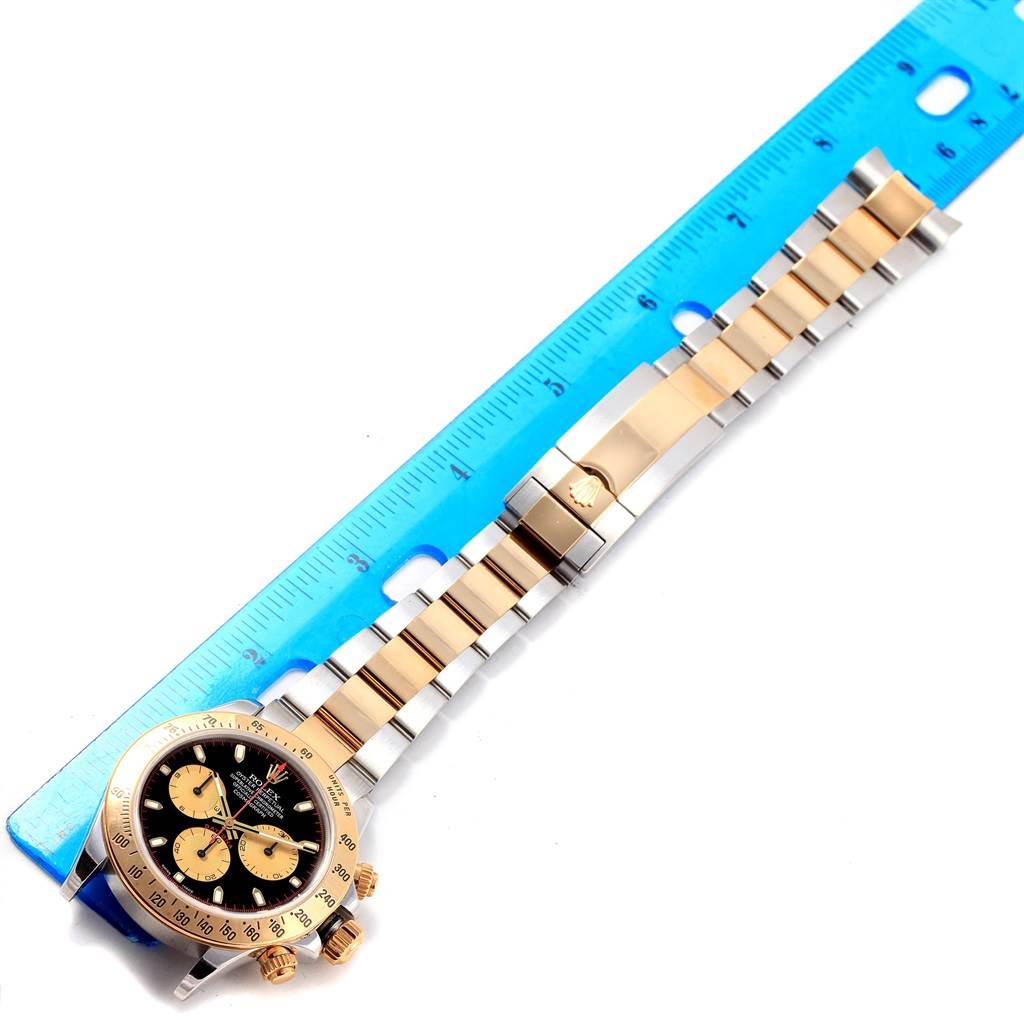 The image shows a Rolex Daytona 116523 Men's Steel and Gold (two tone) Black Dial 116523 Men's Steel and Gold (two tone) Black Dial watch laid flat next to a blue ruler, displaying the dial, case, and bracelet.