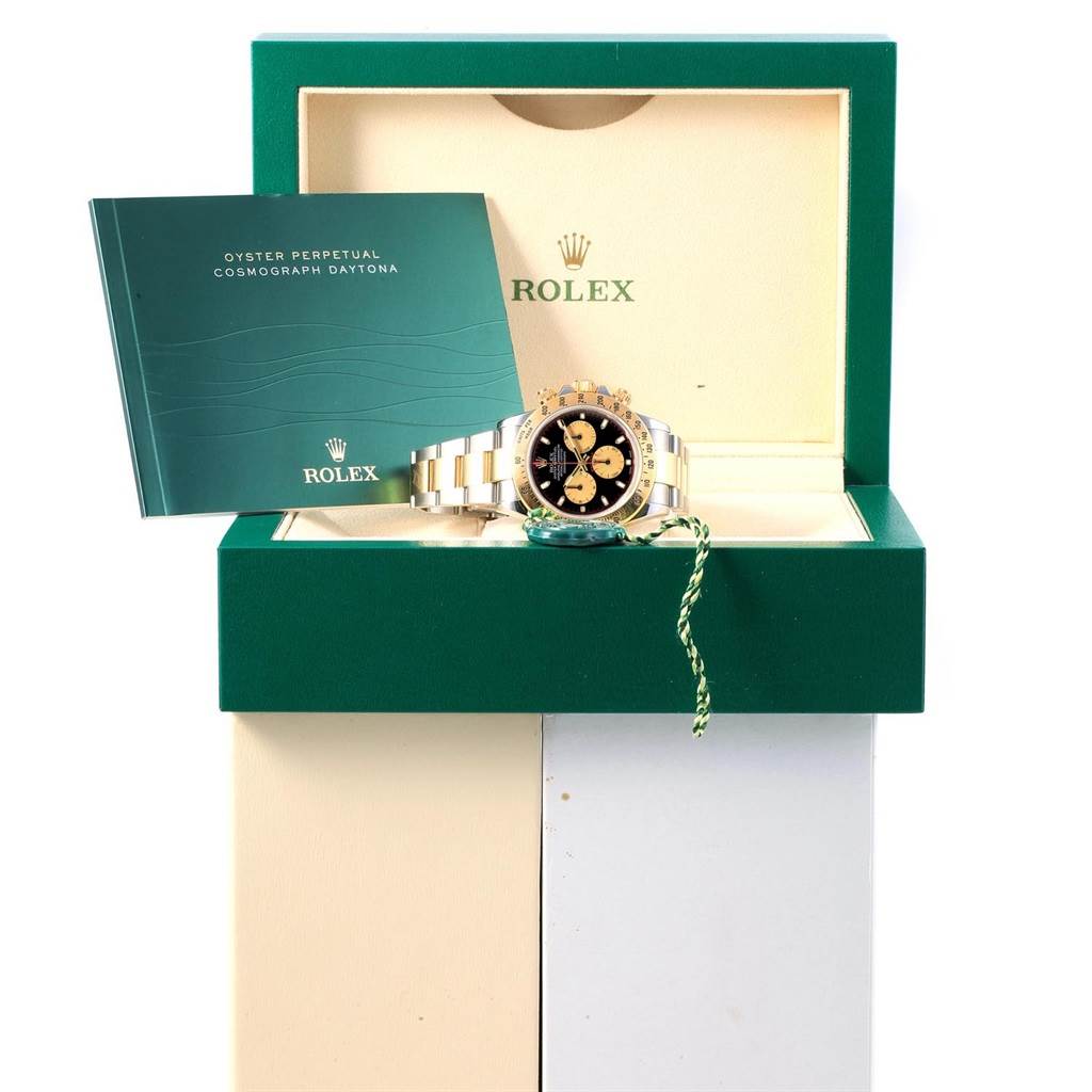 The image shows a Rolex Daytona 116523 Men's Steel and Gold (two tone) Black Dial 116523 Men's Steel and Gold (two tone) Black Dial watch in its box with its booklet, viewed from the front.