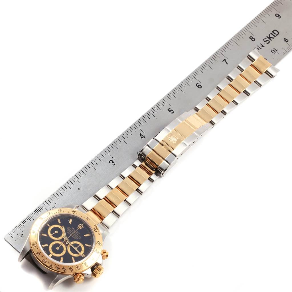 The Rolex Daytona 16523 Men's Steel and Gold (two tone) Black Dial 16523 Men's Steel and Gold (two tone) Black Dial watch is shown from a top-down angle, highlighting its case, bezel, dial, and bracelet alongside a ruler.