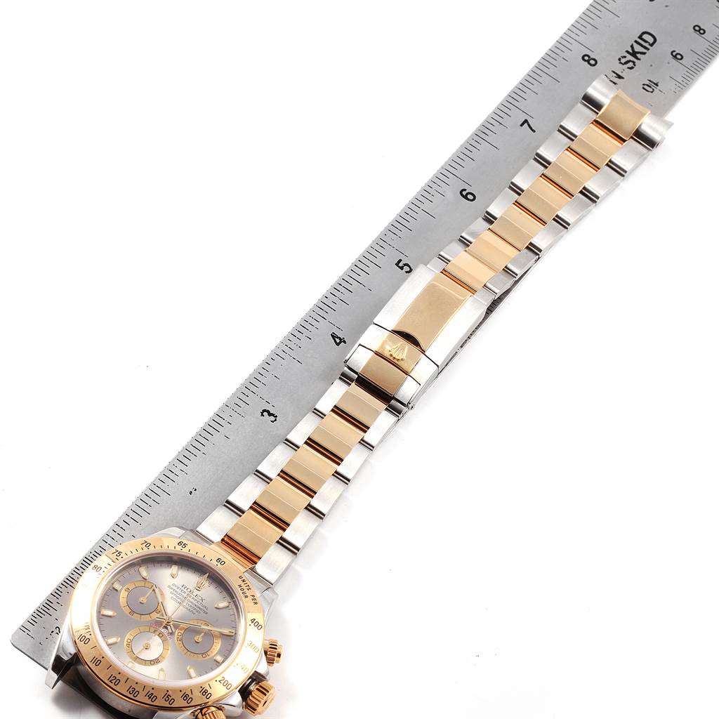 The image shows a Rolex Daytona 116523 Men's Steel and Gold (two tone) Grey Dial 116523 Men's Steel and Gold (two tone) Grey Dial watch laid flat next to a ruler, displaying its bracelet, dial, and case.