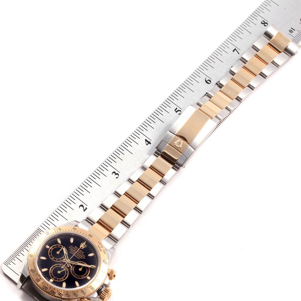 The Rolex Daytona 116523 Men's Steel and Gold (two tone) Black Dial 116523 Men's Steel and Gold (two tone) Black Dial watch is shown from a top view, highlighting its face and bracelet placed along a ruler for scale.