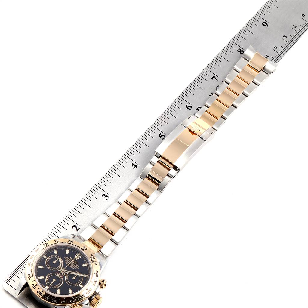 This image shows a Rolex Daytona 116503 Men's Steel and Gold (two tone) Black Dial 116503 Men's Steel and Gold (two tone) Black Dial watch placed next to a ruler, displaying its metallic band and partial view of the dial.