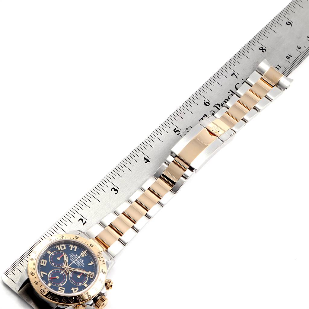 The image shows a Rolex Daytona 116523 Men's Steel and Gold (two tone) Blue Dial 116523 Men's Steel and Gold (two tone) Blue Dial watch laid flat against a ruler, displaying its face and metal bracelet.
