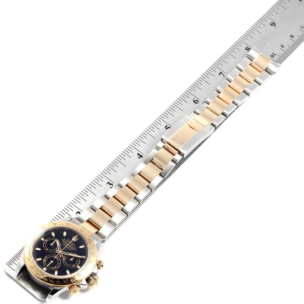 The image shows a Rolex Daytona 116523 Men's Steel and Gold (two tone) Black Dial 116523 Men's Steel and Gold (two tone) Black Dial watch placed against a ruler, highlighting the watch's face, crown, and bracelet.
