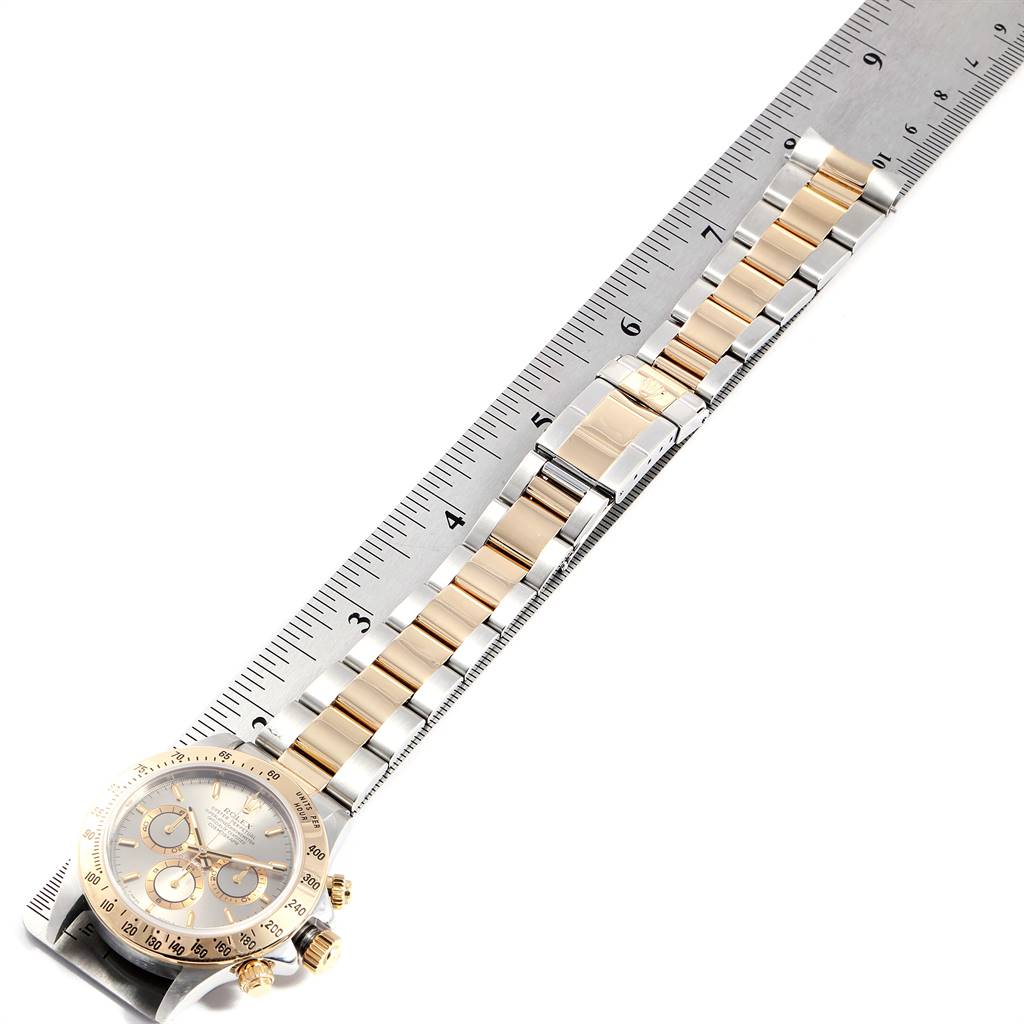 The image shows a front view of a Rolex Daytona 16523 Men's Steel and Gold (two tone) Grey Dial 16523 Men's Steel and Gold (two tone) Grey Dial watch, highlighting its face, crown, and two-tone bracelet, placed alongside a ruler.