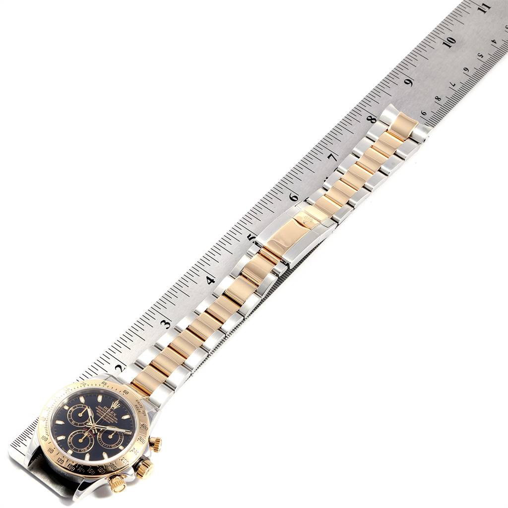 The image shows a Rolex Daytona 116523 Men's Steel and Gold (two tone) Black Dial 116523 Men's Steel and Gold (two tone) Black Dial watch, angled diagonally, with the face and full bracelet against a ruler.