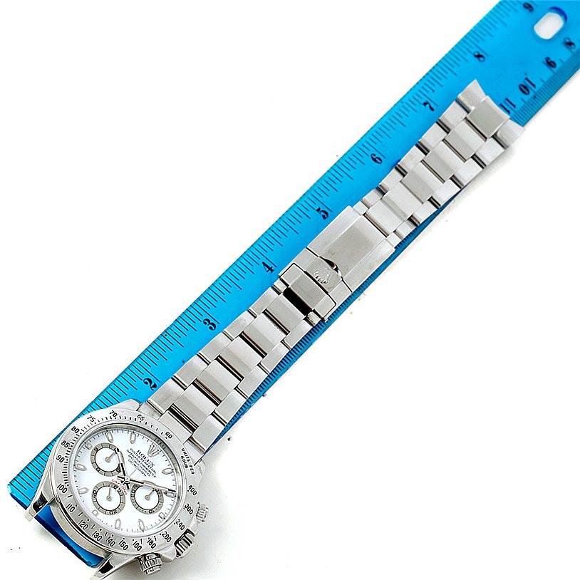The image shows the entire Rolex Daytona 116520 Men
s Stainless Steel White Dial 116520 Men
s Stainless Steel White Dial watch, including the face, bezel, and bracelet, laid next to a blue ruler.