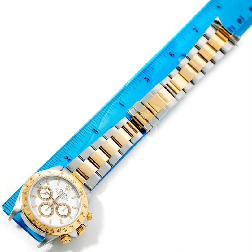 The image shows a Rolex Daytona 16523 Men's Steel and Gold (two tone) White Dial 16523 Men's Steel and Gold (two tone) White Dial watch laid flat next to a blue ruler, displaying its face and bracelet.