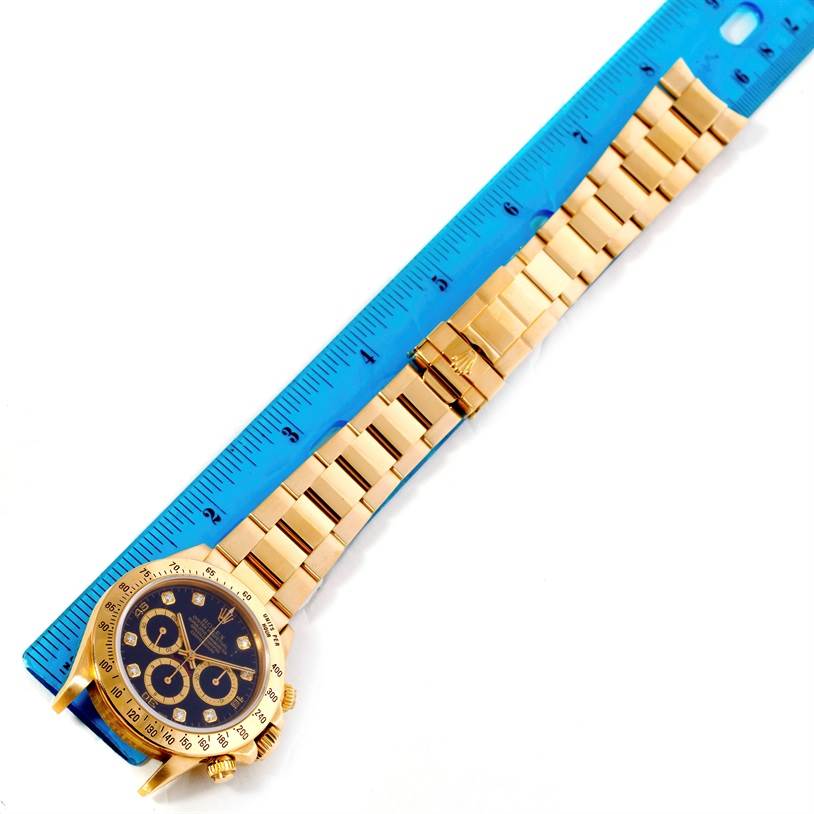 The image shows a top-down view of a Rolex Daytona Men's Yellow Gold Black Dial Men's Yellow Gold Black Dial watch, specifically highlighting the face, bezel, and bracelet next to a blue ruler.